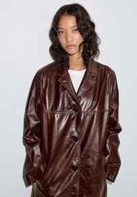 Bruine leren jas met een glanzende afwerking, voorzien van een knoopsluiting, brede revers en zijzakken. Draagt over een wit shirt.