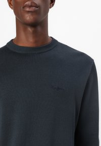 Dunkelblauer Strickpullover mit einem gerippten Rundhalsauschnitt. Verfügt über ein dezentes gesticktes Logo in einem etwas dunkleren Farbton auf der Brust.