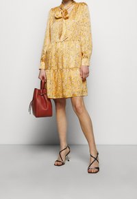Robe jaune à motifs avec des manches longues, un col noué et un design à plusieurs couches. Associée à un sac à main rouge et des talons noirs à lanières.