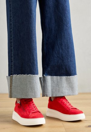 Osoba nosząca szerokie, podwinięte ciemnoniebieskie jeansy i jasnoczerwone trampki na platformie stojąca na drewnianej podłodze.
