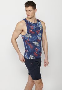 Tengerészi tank top trópusi levél mintával piros, krém és türkiz színekben; kerek nyakkivágással és széles karöltőkkel rendelkezik. Nápoly sorttal párosítva.