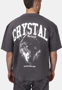 Šedé oversized tričko s grafikou vlka na zádech, s tučným bílým textem "CRYSTAL Paris." Vyrobeno z měkké, texturované bavlny.