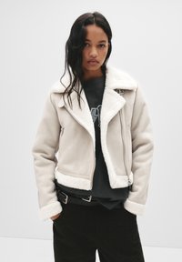 Kremowa kurtka z sztucznego owczej skóry z wyciętym kołnierzem, zamkiem z przodu i bocznymi kieszeniami. Połączona z ciemną koszulą i czarnymi spodniami.