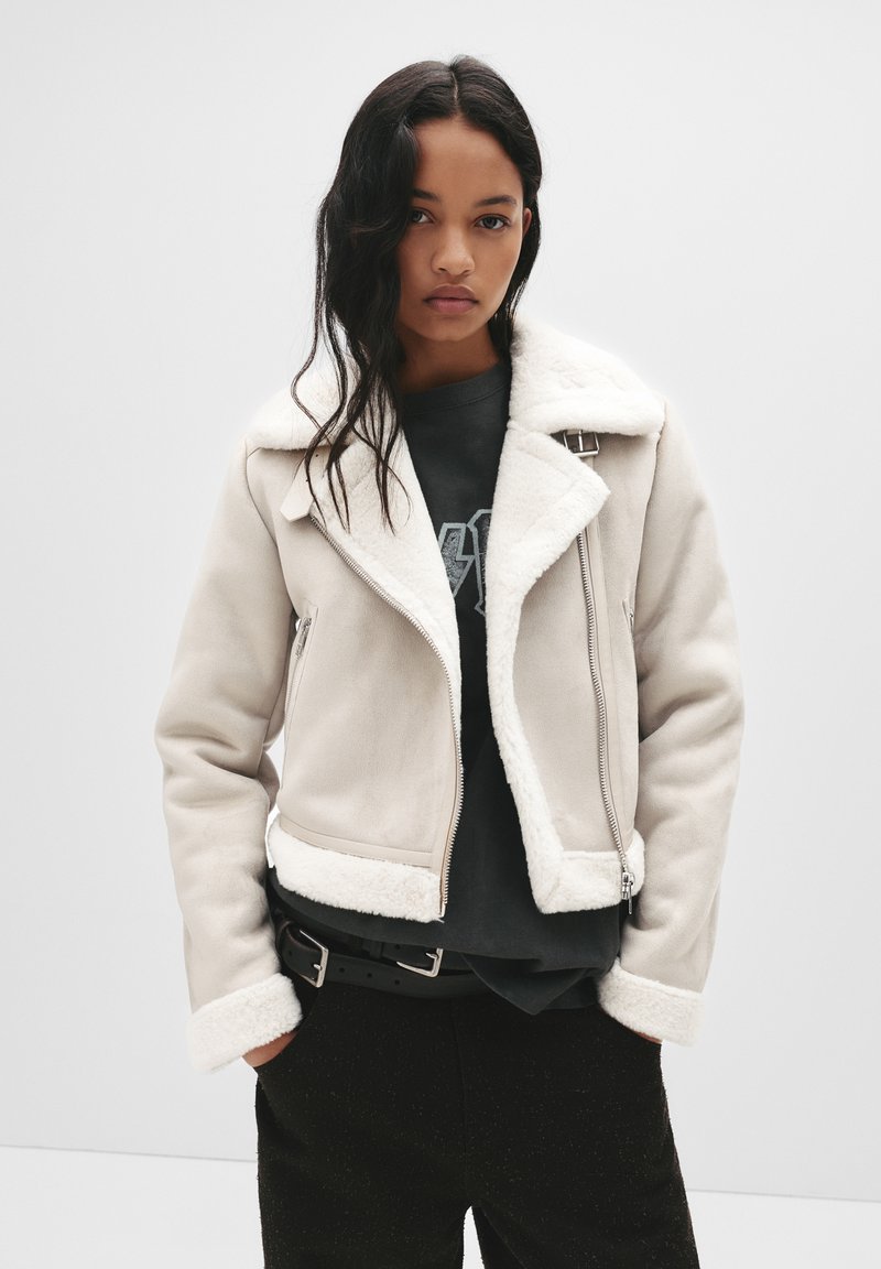 Kremowa kurtka z sztucznego owczej skóry z wyciętym kołnierzem, zamkiem z przodu i bocznymi kieszeniami. Połączona z ciemną koszulą i czarnymi spodniami.