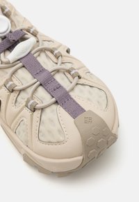 Beige idrettssko med mesh-paneler, lilla aksent-snører og en teksturert gummisåle. Har en avrundet tå og fleksible designelementer.