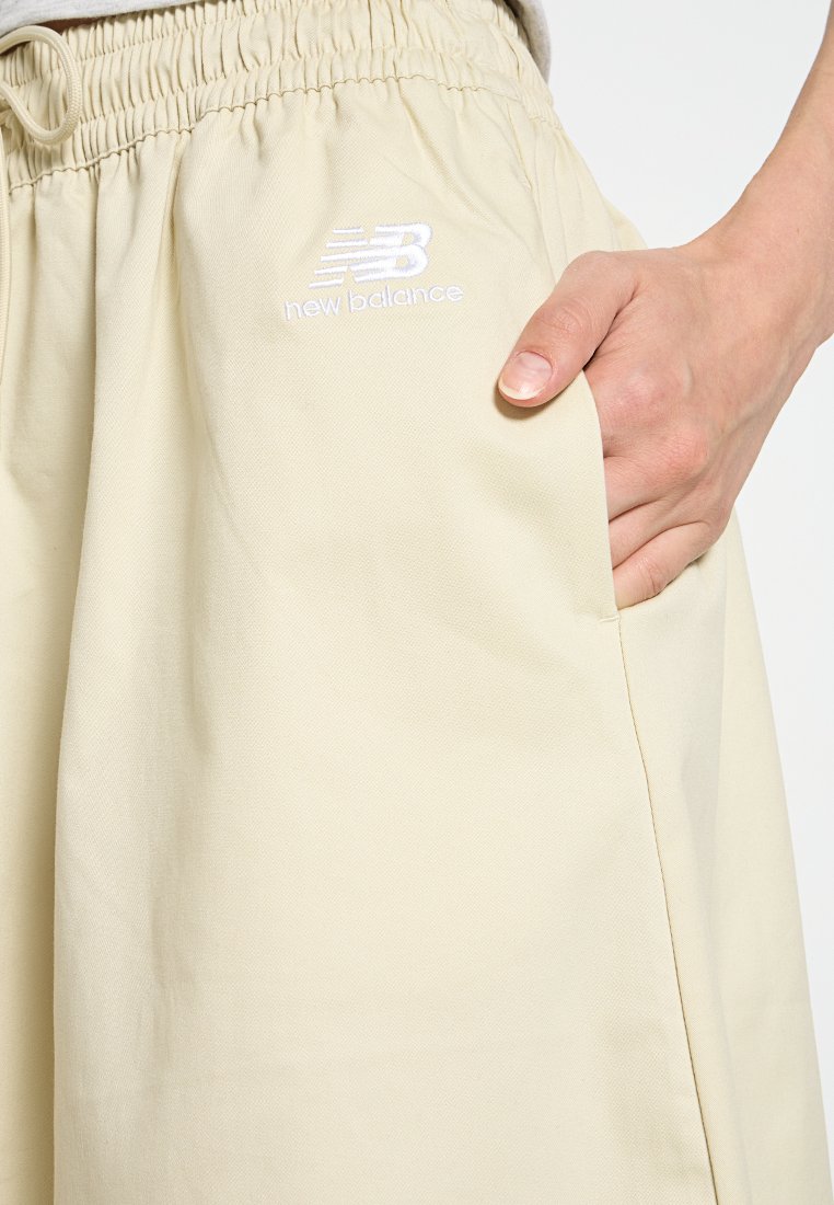 Pantalones de chándal New Balance beige con cinturilla elástica y bolsillo. Presenta un logo bordado en la parte frontal en blanco. Textura de tela suave.