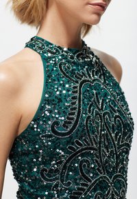 Abito halter neck teal con intricati ricami e paillettes. Decorazioni dettagliate creano un motivo, evidenziando texture e lucentezza.