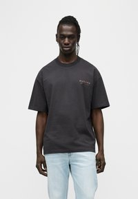 Μαύρο βαμβακερό t-shirt με άνετη εφαρμογή, κοντά μανίκια και μικρή λεπτομέρεια λογότυπου στο στήθος. Συνδυάζεται με ανοιχτόχρωμο τζιν παντελόνι.