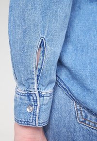 Ljusa blå denimskjortärm med knappstängning och ett utskuret detaljerat mönster. Tyget har en texturerad väv och synliga sömnadsdetaljer.