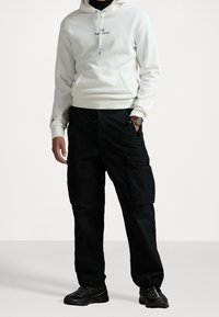 Vit bomullshoodie med framficka och svart logotyp, kombinerad med svarta cargobyxor med sidfickor och elastiska manschetter, bärs med mörka sneakers.