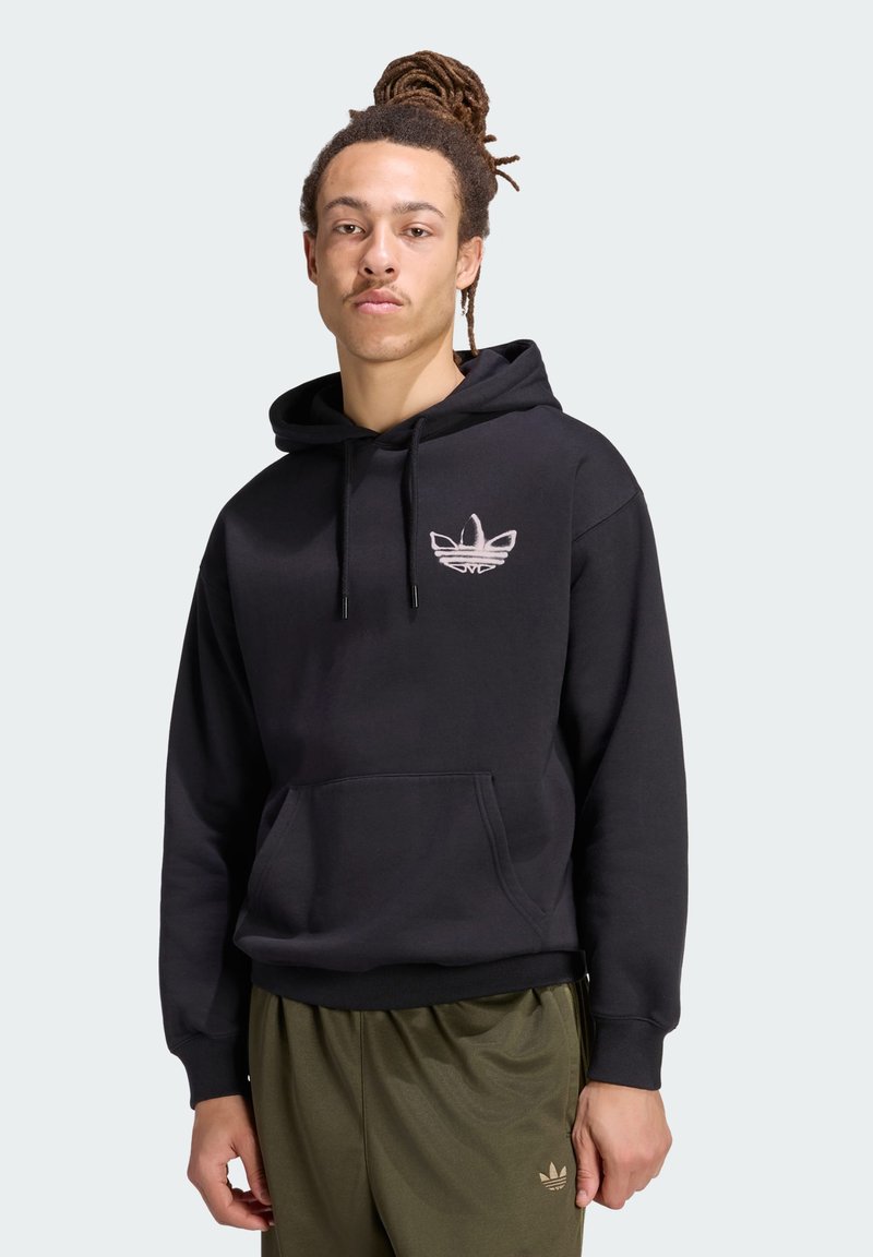 Jeune homme avec dreadlocks portant un sweat à capuche noir et un pantalon vert olive, debout devant un fond clair uni.