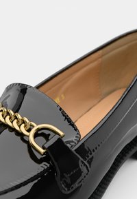 Mocassini in pelle verniciata nera con accentuazione in catena dorata e bordi testurizzati; l'interno è beige con cuciture minime.