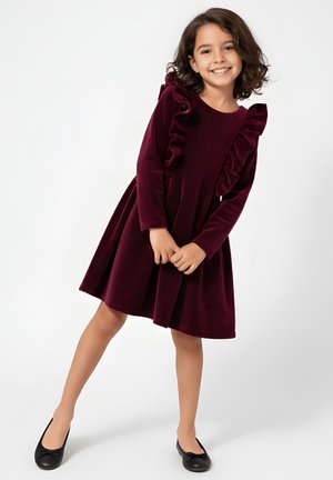 Bordowa aksamitna sukienka z długimi rękawami i falbanami na ramionach; rozkloszowana spódnica, skomponowana z czarnymi balerinami.