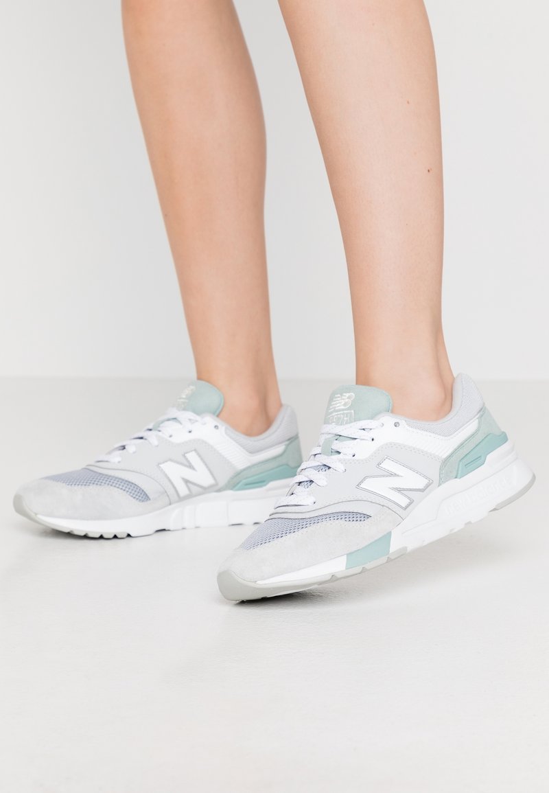 Paire de jambes portant des baskets New Balance blanches et bleu clair, debout sur une surface blanche.