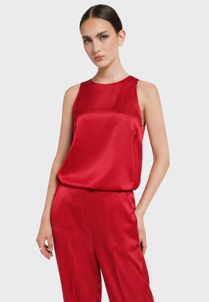 Kobieta o ciemnych włosach ubrana w czerwoną, jedwabistą bluzkę bez rękawów oraz pasujące szerokie spodnie, stojąca na tle jasnego, jednolitego tła.