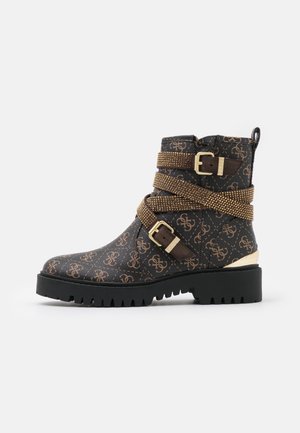Bottine marron foncé avec imprimé monogramme beige, deux sangles cloutées dorées avec boucles, semelle à crampons noire et accent talon doré.
