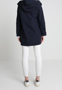 Cazadora impermeable azul marino con capucha, dos botones en el cuello y mangas ajustables. Combinada con jeans ajustados blancos y zapatillas beige.