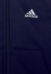 Donkerblauwe hoodie met ritssluiting met een gladde textuur, voorzien van een wit Adidas-logo en zijzakken. De stof lijkt zacht en ademend.