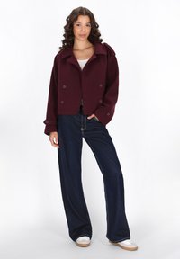 Junge Frau mit lockigem Haar, die eine bordeauxrote, doppelt geknöpfte Jacke, dunkle weite Jeans, weiße Sneakers und ein weißes Oberteil trägt, steht.