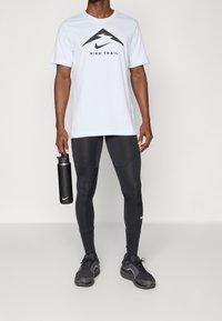 Světle modré krátké tričko značky Nike s černým logem, v kombinaci s černými elastickými legínami a černými sportovními botami. V ruce drží láhev s vodou.