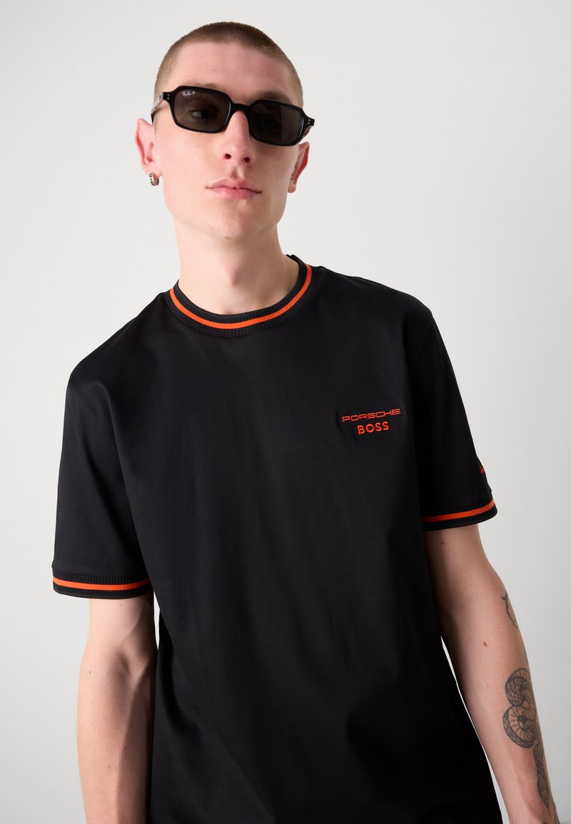 T-shirt noir à manches courtes avec un col rond et des accents orange, un logo sur la poitrine portant "Porsche BOSS", et une texture lisse.