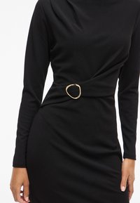 Rochie neagră cu mâneci lungi, cu un detaliu răsucit la talie fixat de o cataramă neregulată aurie, purtată de o persoană cu mâinile relaxate pe lângă corp.