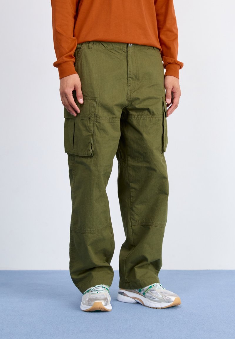 Une personne porte un pantalon cargo vert olive avec de grandes poches latérales, des baskets blanches et vertes, et un haut à manches longues orange, debout sur un sol bleu.