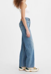 Femme en jean bleu clair à jambes larges, débardeur blanc et baskets beiges, debout de profil sur fond blanc.