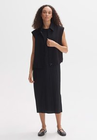 Black sleeveless vest over a pleated black midi dress, featuring a structured collar and lightweight fabric. Model wears black flats.