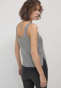 Jeune femme aux cheveux bouclés portant un débardeur en tricot gris sans manches et un jean noir, montrant un tatouage représentant le visage d'un chien sur le haut de son dos.
