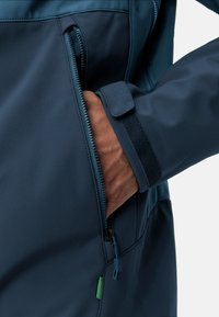 Veste imperméable marine avec une poche zippée, un tissu tactile, des poignets ajustables avec Velcro et un détail de fermeture éclair contrastant.