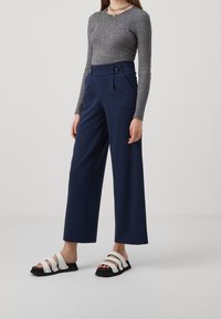 Marinblå byxor med vida ben, framvikta veck, hög midja och knappar. Matchade med en grå ribbad långärmad topp och svartvita sandaler.