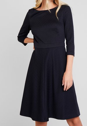 Marineblauwe jurk met een gestructureerd diamantpatroon, driekwartmouwen, een ronde halslijn en een getailleerde taille die uitloopt in een wijd uitlopende rok.