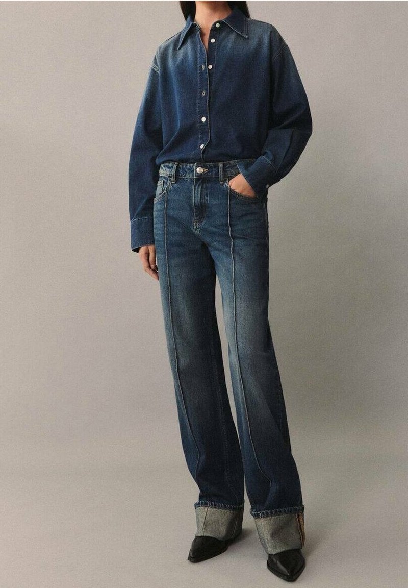 Personne portant une chemise en jean bleu foncé rentrée dans un jean taille haute avec de larges revers et des chaussures noires à bout pointu, debout devant un fond uni.