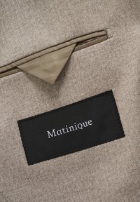 Beiger, strukturierter Stoff mit einem kleinen schwarzen Etikett, auf dem in weißer Schrift "Matinique" steht. Enthält eine kontrastierende braune Stofftasche.