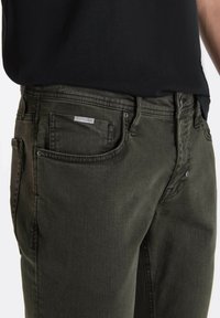 Jeans de mezclilla verde oliva con corte ajustado, que cuentan con cinco bolsillos, botones de metal y detalles de costura. Etiqueta visible encima del bolsillo trasero.