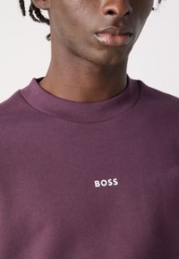 Sudadera morada con cuello redondo acanalado y un pequeño logo blanco "BOSS" en el pecho. Textura suave, diseño ceñido.