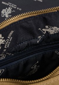 Vaaleanruskea vetoketjullinen pussi, jonka sininen sisusta on koristeltu toistuvalla vaaleanharmaalla hevosen polon kuviolla ja jonka saumanvarassa on sininen ommel.