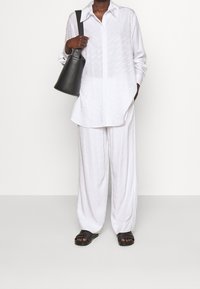 Persona che indossa una camicia bianca larga e pantaloni, sandali neri e porta una borsa a tracolla nera, in piedi su uno sfondo semplice.