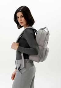 Zaino grigio di forma rettangolare, realizzato in materiale liscio. Presenta spallacci regolabili, tasche laterali e un compartimento frontale con zip.