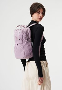 Lila Nylon-Rucksack mit doppelten Obergriffen, verstellbaren Trägern und dezenten kreisförmigen Akzenten. Glatte Textur mit Detail auf der Vorderseite.