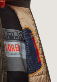 Pufferjakke i olivengrøn med en beige foring, der har en indvendig navy-lomme med mesh-struktur og en kontrasterende rød lynlås.