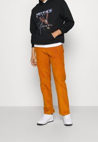 Sudadera negra con estampado gráfico, pantalones de pana naranja y zapatillas blancas. El conjunto presenta un corte relajado y una paleta de colores contrastantes.
