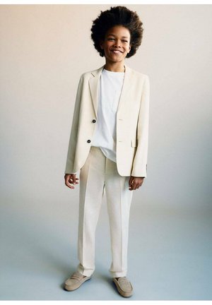 Completo beige chiaro con giacca su misura e pantaloni dritti, abbinato a una camicia bianca e scarpe slip-on beige. Presenta un tessuto testurizzato.