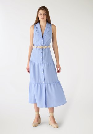 Donna in piedi che indossa un vestito azzurro chiaro senza maniche con bottoni, una cintura intrecciata beige e zeppa beige con suola in corda.