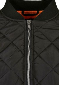 Veste matelassée noire avec fermeture éclair argentée et col côtelé noir, laissant apparaître un soupçon de tissu orange à l’intérieur de l’encolure.