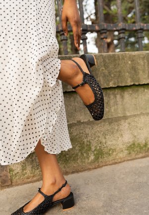 Person, die einen schwarzen gewebten Schuh mit niedrigem Absatz anpasst, trägt ein weißes Midikleid mit schwarzen Punkten und steht im Freien nahe einer Steinmauer und einem Eisenzaun.