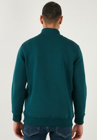 Felpa color teal con collo alto, maniche lunghe e polsini e orlo a costine. Caratterizzata da una texture liscia e una silhouette aderente.