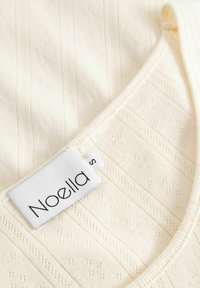Ljus krämfärgad topp med subtila vertikala ränder och texturerade prickar. Har en rundad halsringning och en storleksetikett med "Noella".