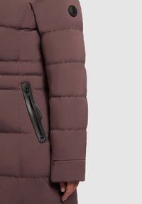 Pufi kabát tompított mályvás színben, vízszintes varrásokkal, oldalsó cipzáros zsebbel és fekete logóval a váll közelében. Simább, texturált anyag.